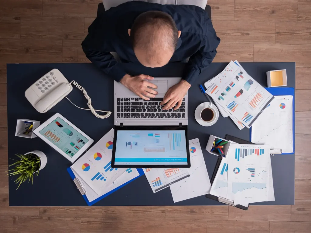 Top down view of man analysing papers and laptop at his desk trying to process manual information